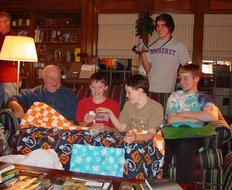 Willie sits on the sofa and receives a quilt, while Grandpa, Andy, and Phil seated with him look on, and behind them Scott has a video camera
